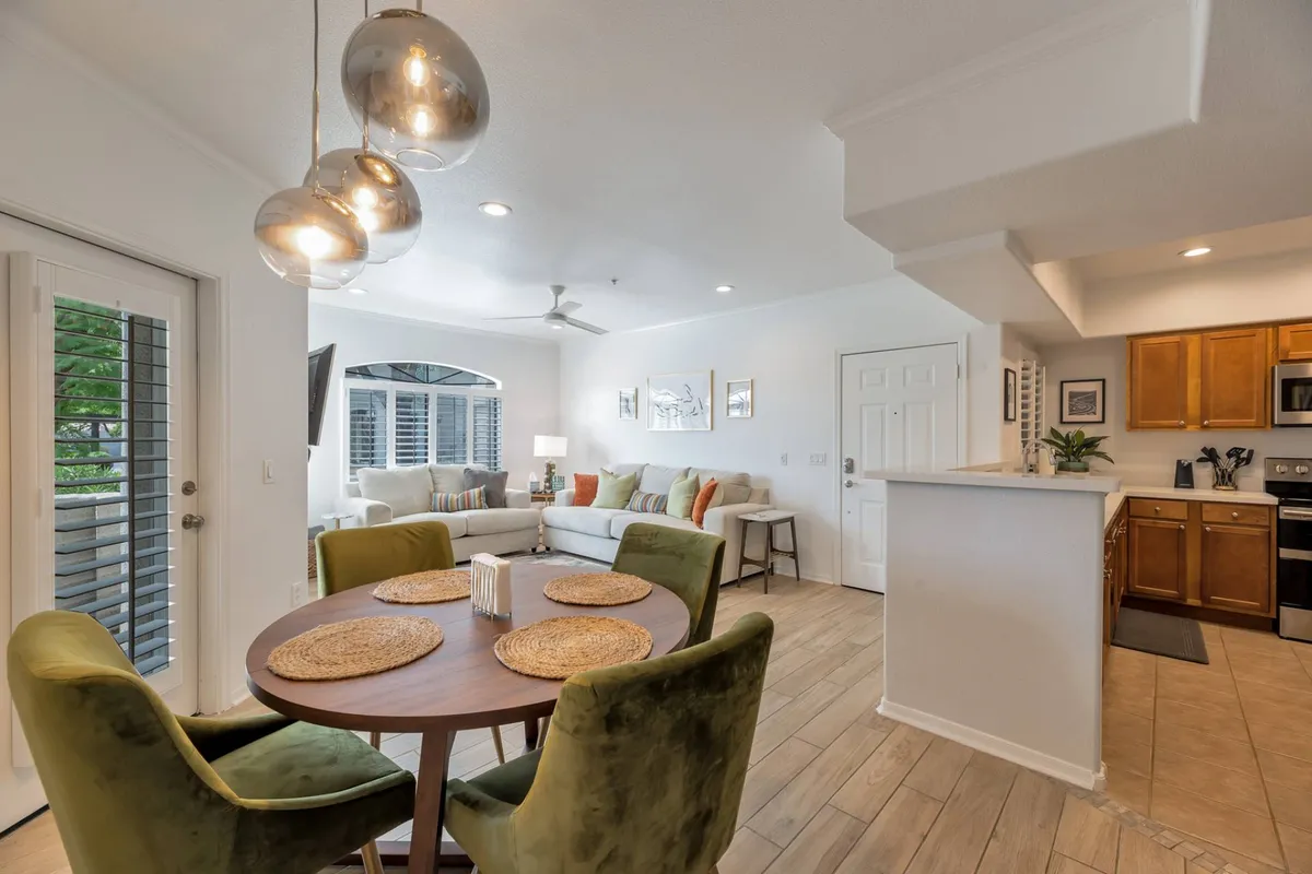 An inviting open-layout scene that features a round dining table for four with green chairs, a sofa in the background, window blinds, and kitchen appliances nearby. The arrangement encourages socializing and relaxed meals in a connected space.