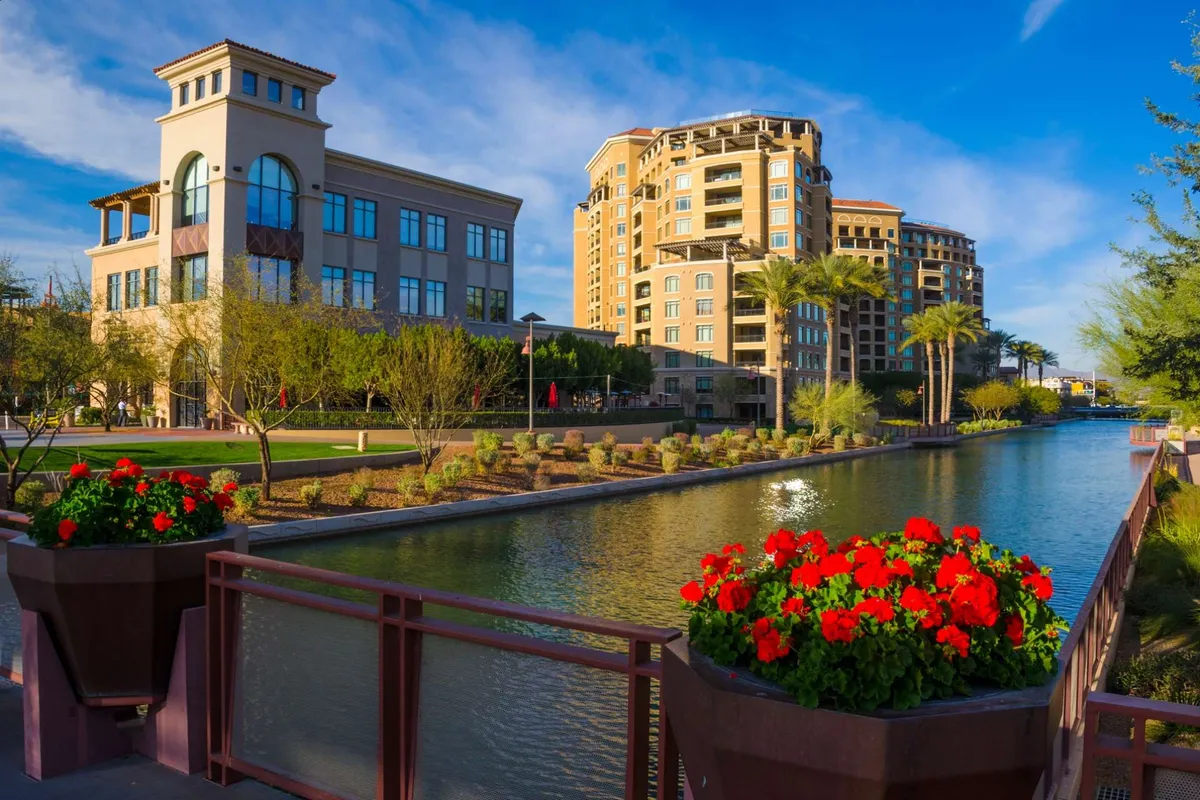 A picturesque exterior with modern buildings, lush landscaping, vibrant flower planters, and calm water reflecting the surroundings. People are seen enjoying the community space, walking by the waterway, relaxing, and exploring shops, illustrating the lively atmosphere of Scottsdale’s shopping and dining districts.