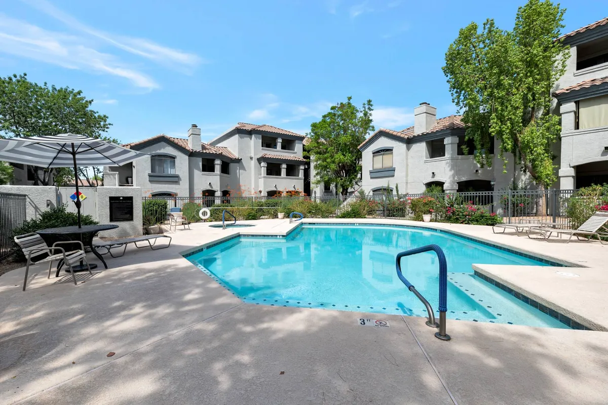 A sunlit exterior view of the community's heated swimming pool surrounded by trees and inviting patio furniture. The pool area is adjacent to welcoming building architecture, signaling a family-friendly and relaxing environment perfect for cooling off after a day of adventure.