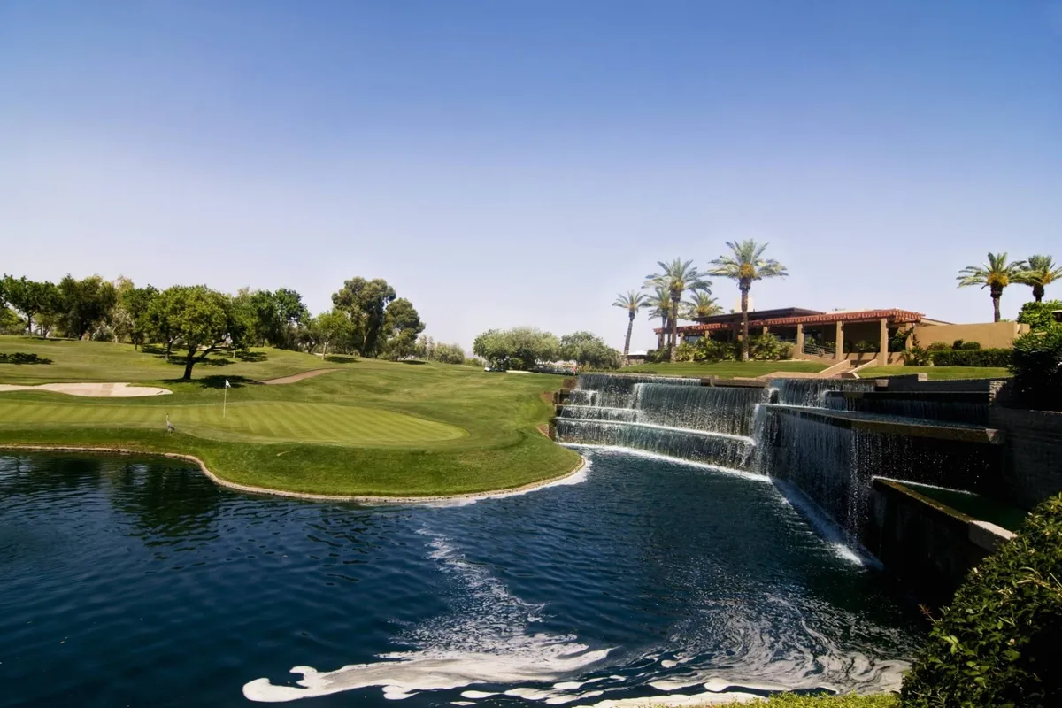 A lush view of the Starfire Golf Club, featuring green fairways, a tranquil waterfall, tall palm trees, and the clubhouse in the background. The scene is inviting for both relaxation and recreation, with the golf course as the focal point.