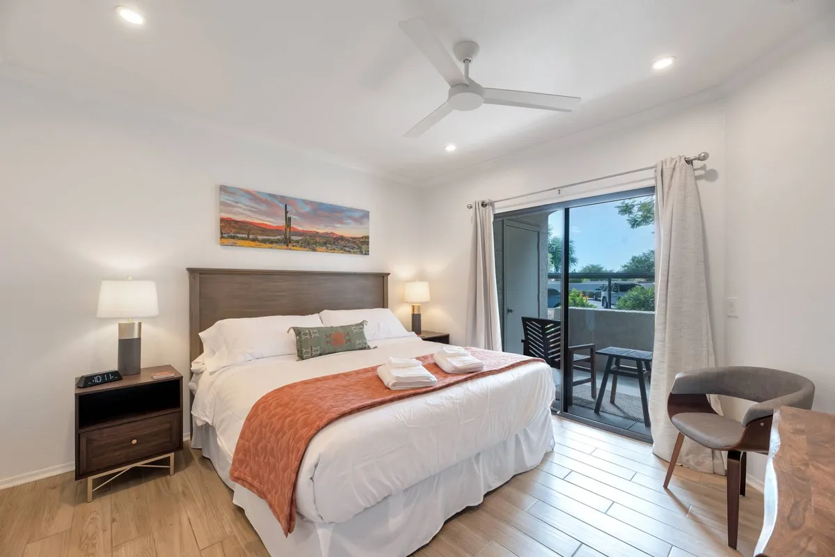 This image showcases a spacious bedroom with crisp white linens, an inviting orange throw blanket, and modern artwork above the bed. A glass door opens directly onto a private patio, allowing for natural light to pour in and offering easy access to fresh desert air. The scene is calm and uncluttered, emphasizing relaxation and tranquility.