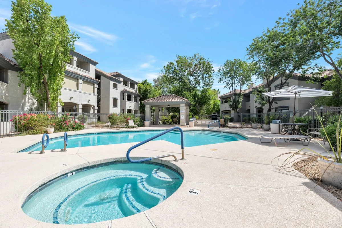 This exterior image showcases a sparkling swimming pool with an adjacent hot tub, surrounded by a beautifully landscaped garden, sun umbrellas, and ample seating areas. The relaxed atmosphere is perfect for both social gatherings and quiet relaxation, echoing the resort-style theme of the amenities.